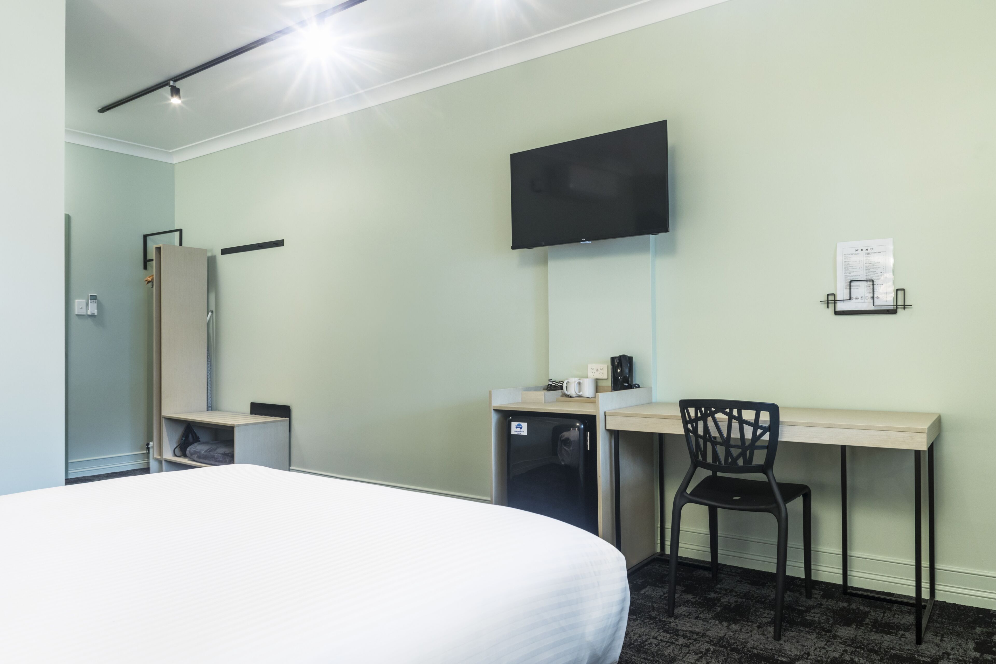 A hotel room with a bed, a desk with a chair, a television mounted on the wall, and a small refrigerator.
