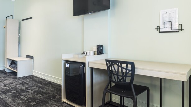 A compact hotel room with a kitchenette, a flat-screen TV mounted on the wall, a small refrigerator, a desk with a chair, and a wall-mounted menu holder. - Nightcap at Atherton Hotel (Atherton)