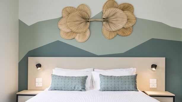 Cama bem arrumada com almofadas estampadas, luminária fixada na parede e um objeto decorativo na parede.