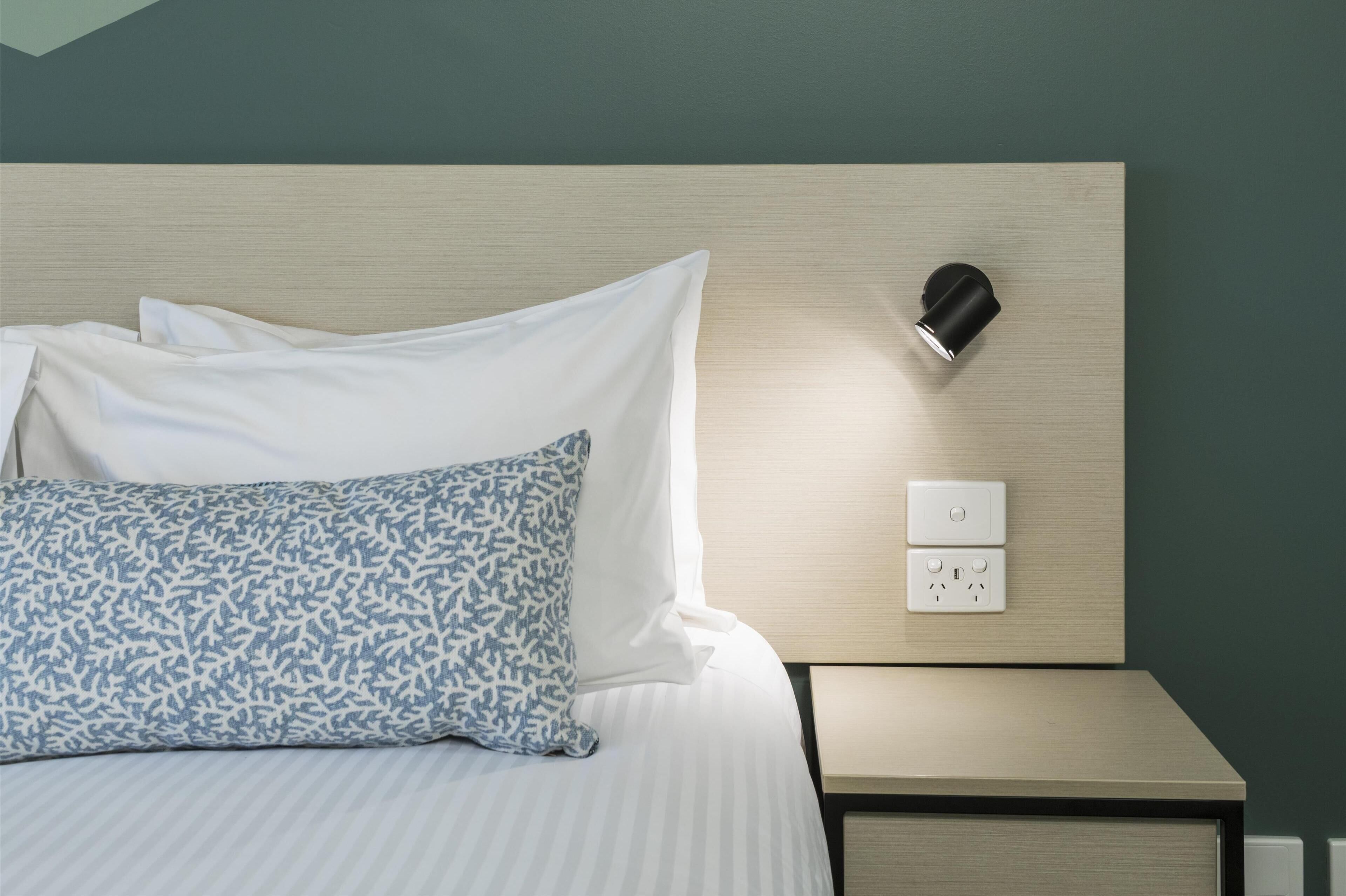 A neatly made bed with white and blue patterned pillows, a bedside table with a lamp, and a wall socket.