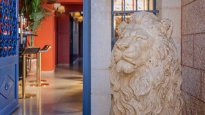Une statue de lion en pierre se trouve dans un couloir, sur un mur orné d’un motif bleu, et un palmier est visible en arrière-plan.