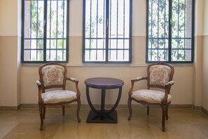 Ein Zimmer mit zwei verzierten Sesseln und einem kleinen runden Tisch, der zwischen zwei vergitterten Fenstern platziert ist.