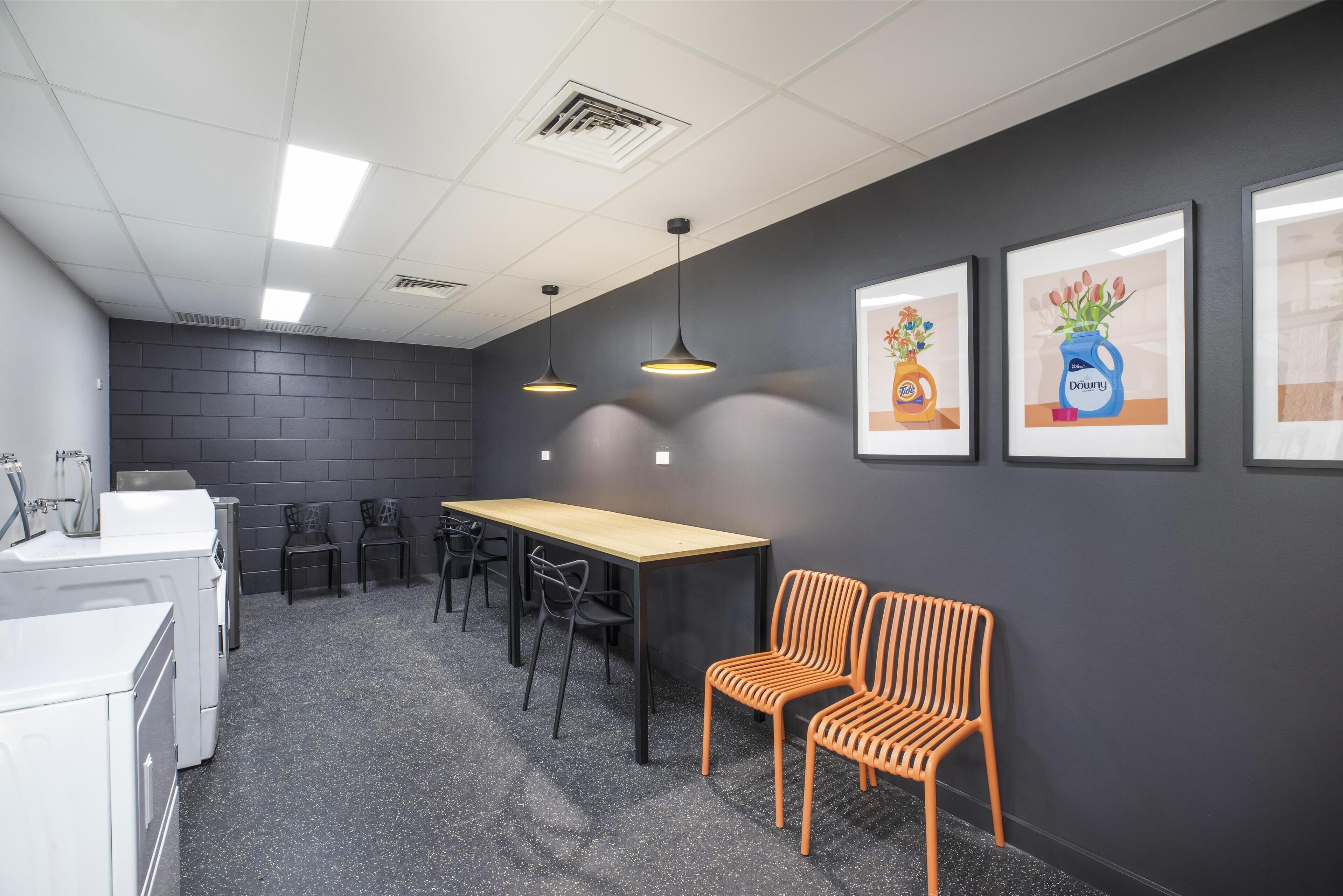 Een vergaderruimte met een lange tafel, stoelen, en twee oranje fauteuils. Er hangen ingelijste foto's aan de muur en staat een wasmachine in de hoek.