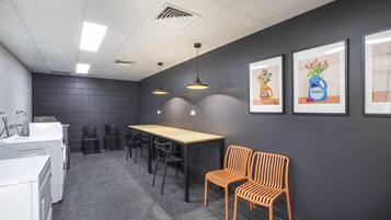 Een vergaderruimte met een lange tafel, stoelen, en twee oranje fauteuils. Er hangen ingelijste foto's aan de muur en staat een wasmachine in de hoek.