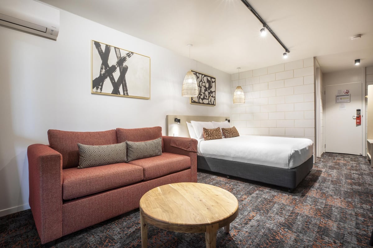 Ein Hotelzimmer mit einem roten Sofa, einem runden Holztisch, einem Bett mit weißer Bettwäsche und zwei an der Wand befestigten Pendelleuchten.