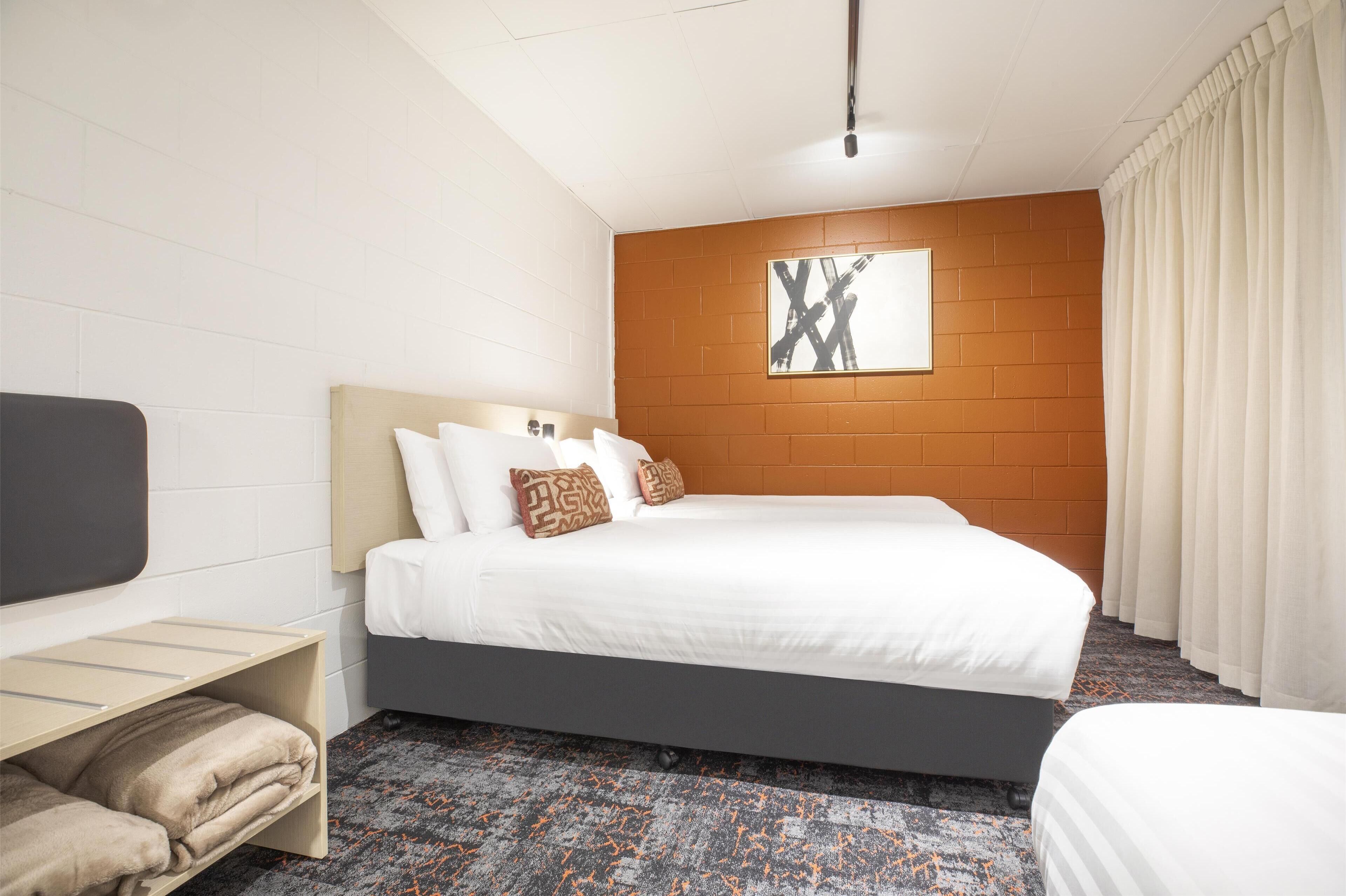 Una habitación de hotel con una cama grande, cabecero, mesita de noche, estante con toallas y alfombra estampada.