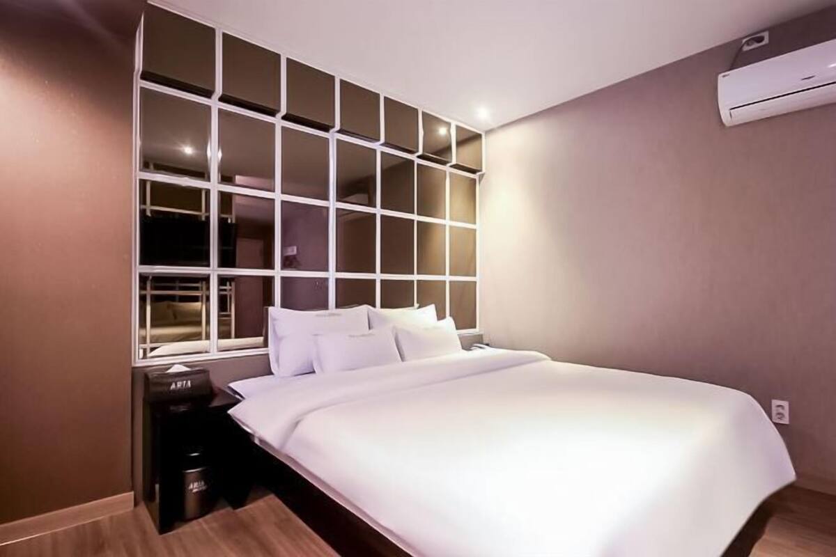 A bedroom with a large window, a bed, a nightstand, and an air conditioning unit.