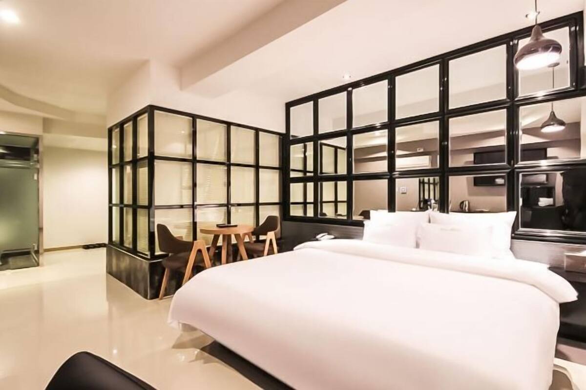 A modern hotel room with a large bed, a seating area with chairs, a glass partition, and a large mirror.