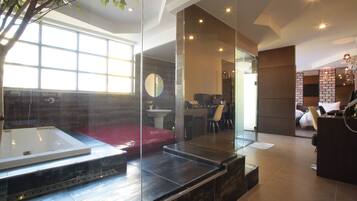 A modern hotel room with a large glass wall, a bathtub, a red sofa, a desk, and a brick accent wall.
