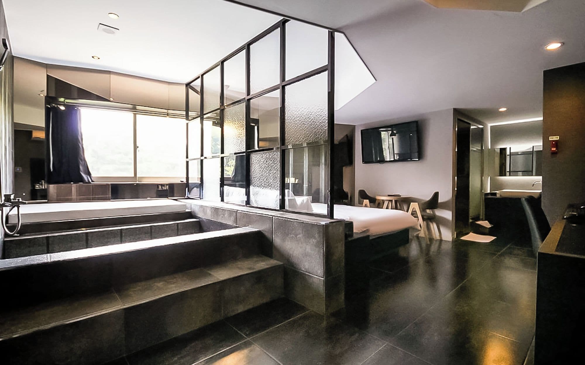 A modern hotel room with a large window, a staircase, a bed, a desk, and a bathroom.