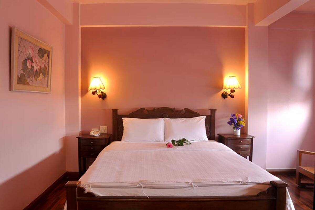 A neatly made bed with white linens, two bedside lamps, a nightstand with a telephone, and a framed floral artwork on the wall.