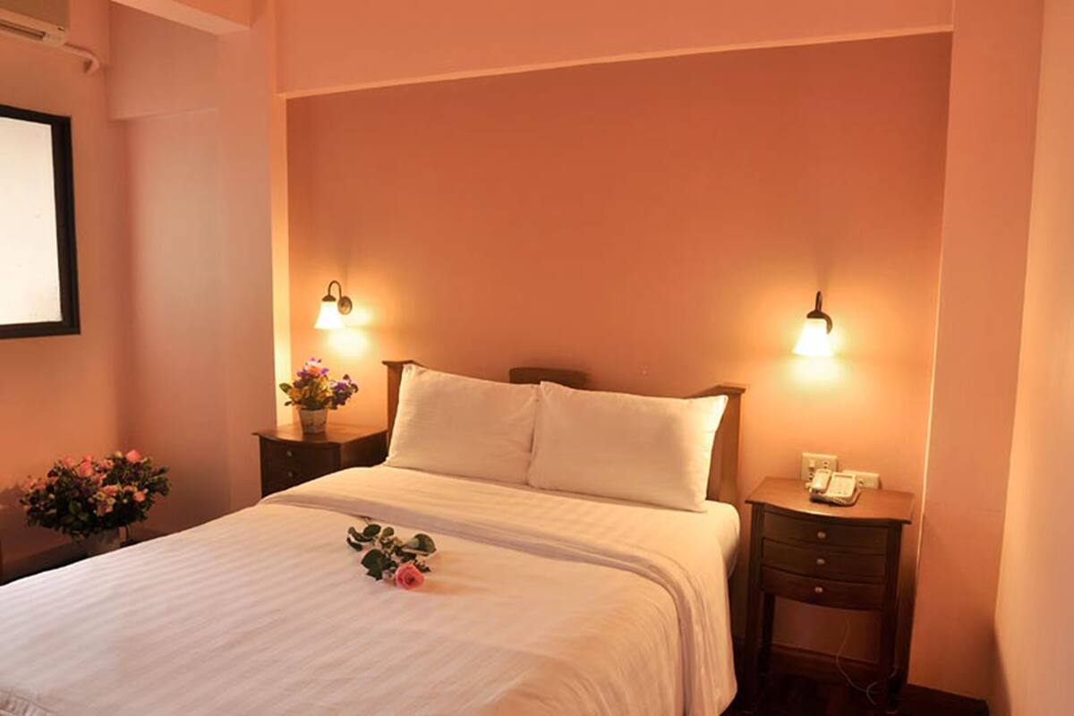 A neatly made bed with white linens and a single rose on the bedspread. There are two bedside tables with lamps, one on each side of the bed.