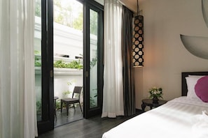 Habitación de hotel con cama, una pequeña mesa con una planta, lámpara colgante y puerta corrediza de vidrio que da a una zona de estar exterior.