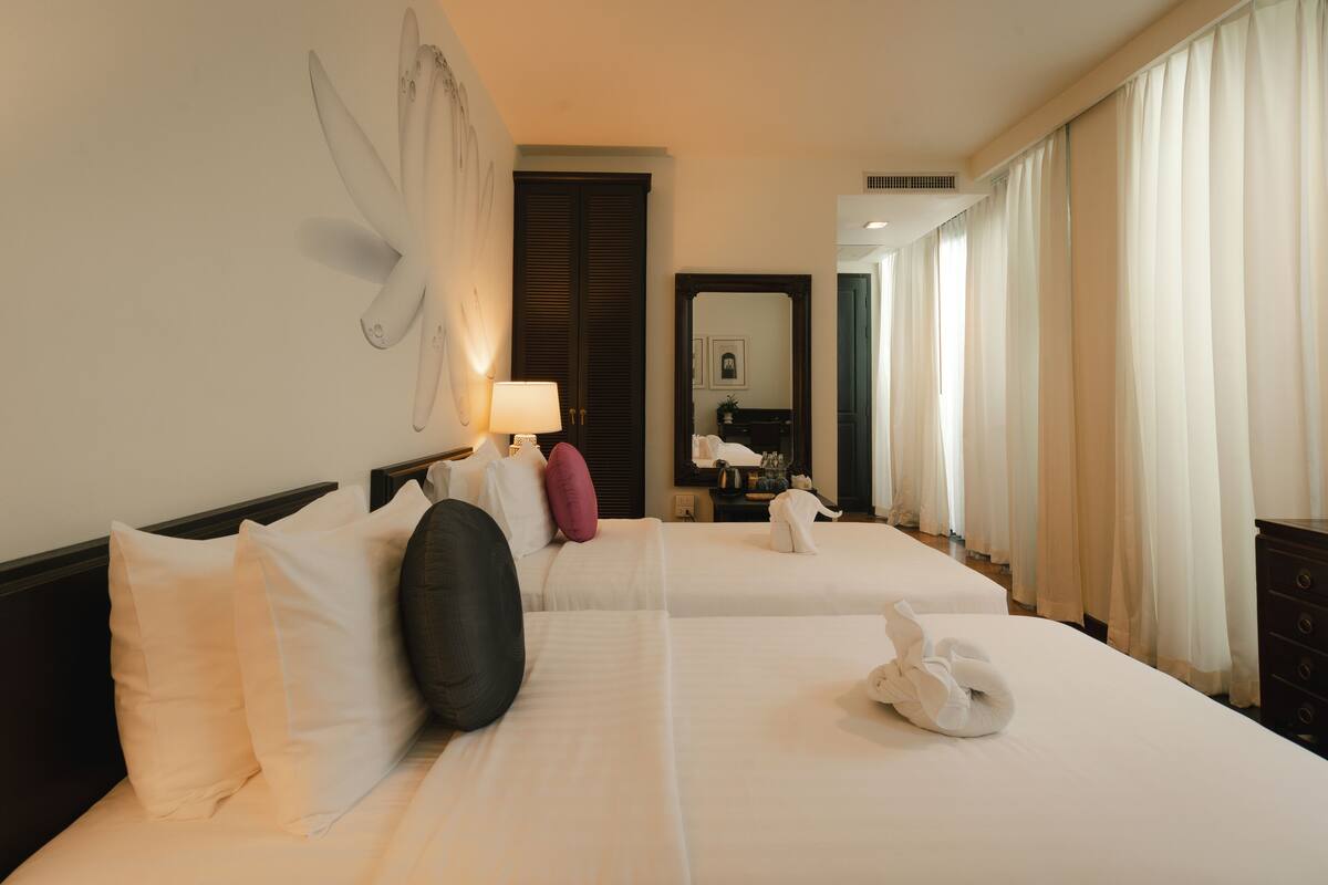 A hotel room with two beds, a large mirror, a wall-mounted lamp, and a window with curtains.