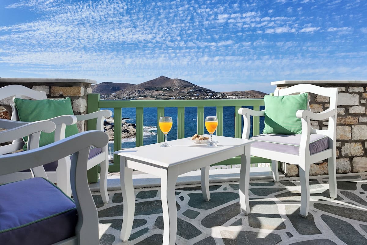 A terrace with white furniture, a table with a glass of orange juice, and a view of the sea and mountains.
