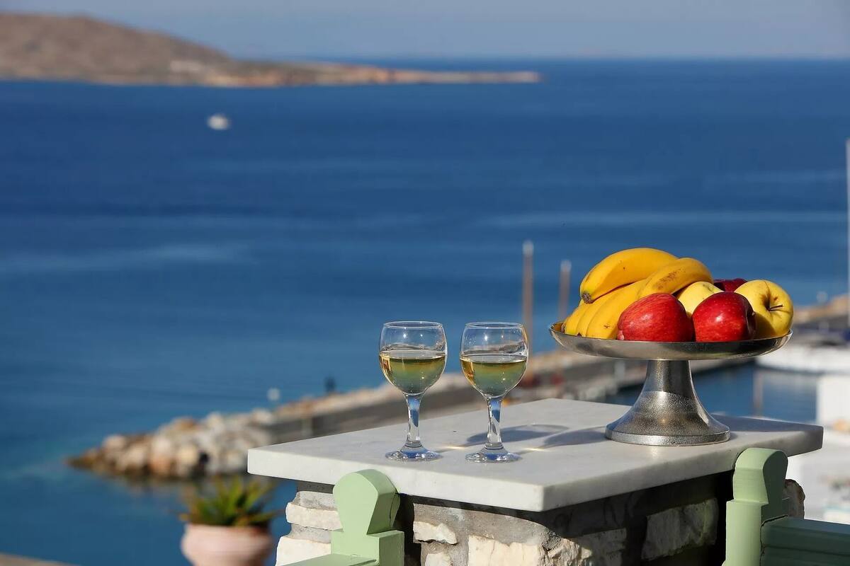 A table with two glasses of white wine, a metal fruit bowl with bananas and apples, and a view of the sea.