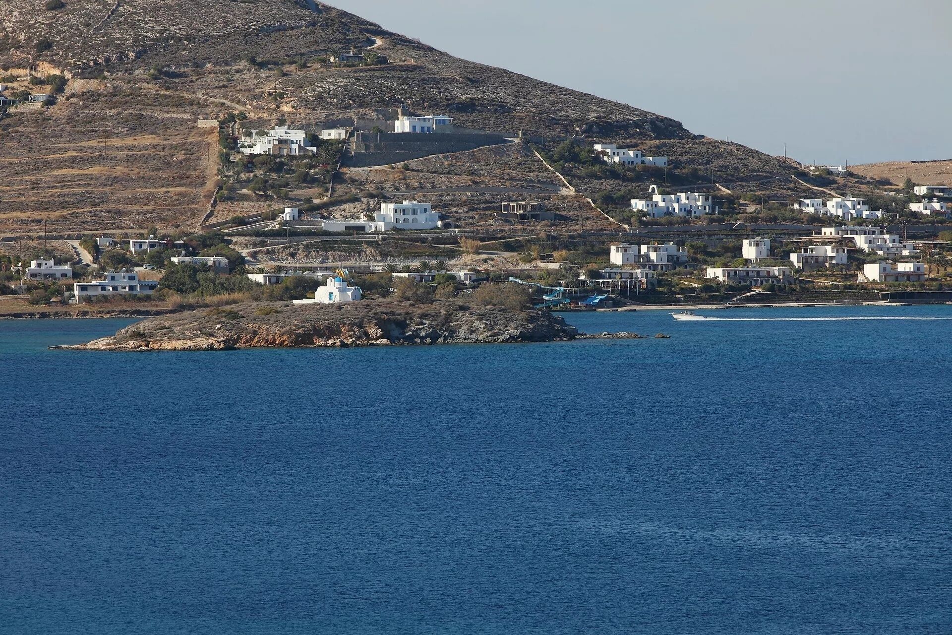 A coastal village with white buildings, a rocky island, and a boat in the water.
