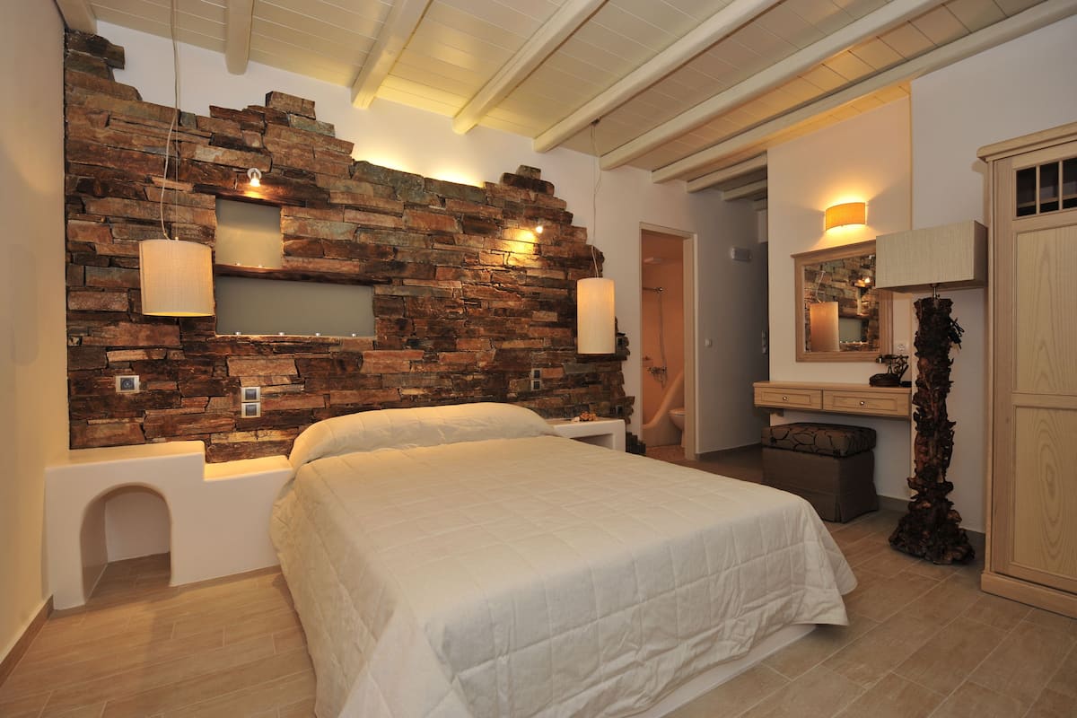 A bedroom with a stone wall, a bed, bedside lamps, a wooden cabinet, and a mirror.