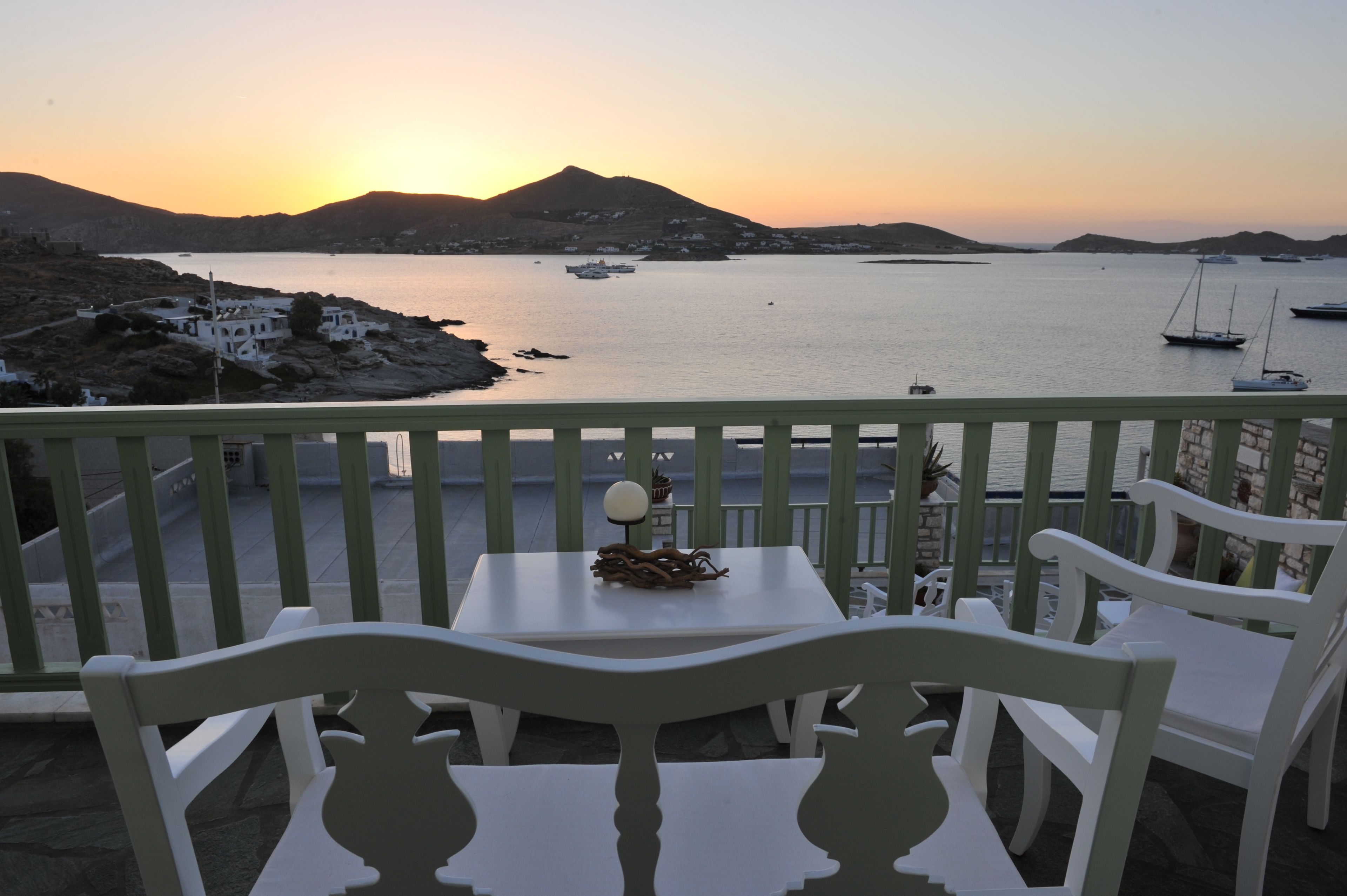A balcony with white chairs and a table, overlooking a calm sea with boats and a mountainous landscape during sunset.