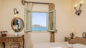 A bedroom with a bed, a desk, a chair, a bedside table with a lamp, a wall-mounted mirror, and a window with curtains. - Villa Isabella Studios & Suites (Paros)