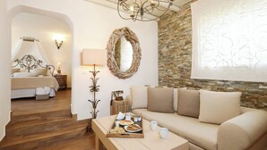 A cozy room with a bed, a sofa, a dining table with breakfast, a chandelier, and a stone wall. - Villa Isabella Studios & Suites (Paros)