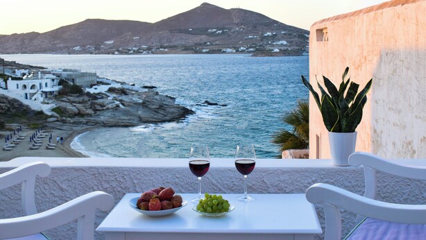 A balcony with a table set for two, featuring a bowl of fruit and two glasses of red wine, overlooking a coastal landscape with a beach and mountains. - Villa Isabella Studios & Suites (Paros)