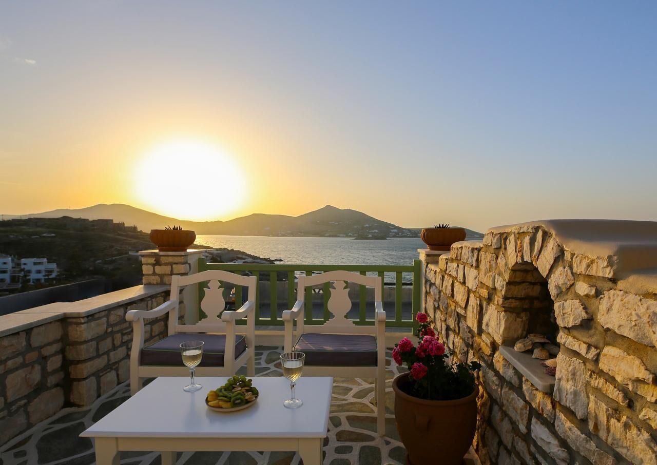 A terrace with a table set for two, a stone wall, and a view of the sea at sunset.