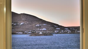 A coastal view through a window, featuring a hillside with buildings and a body of water. - Villa Isabella Studios & Suites (Paros)