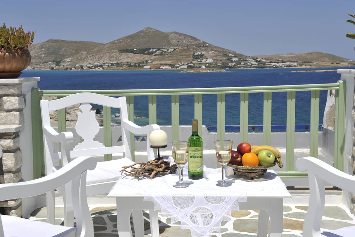 A table with a bottle of wine, glasses, and a bowl of fruit, set on a balcony with a view of the sea and a mountain in the background.