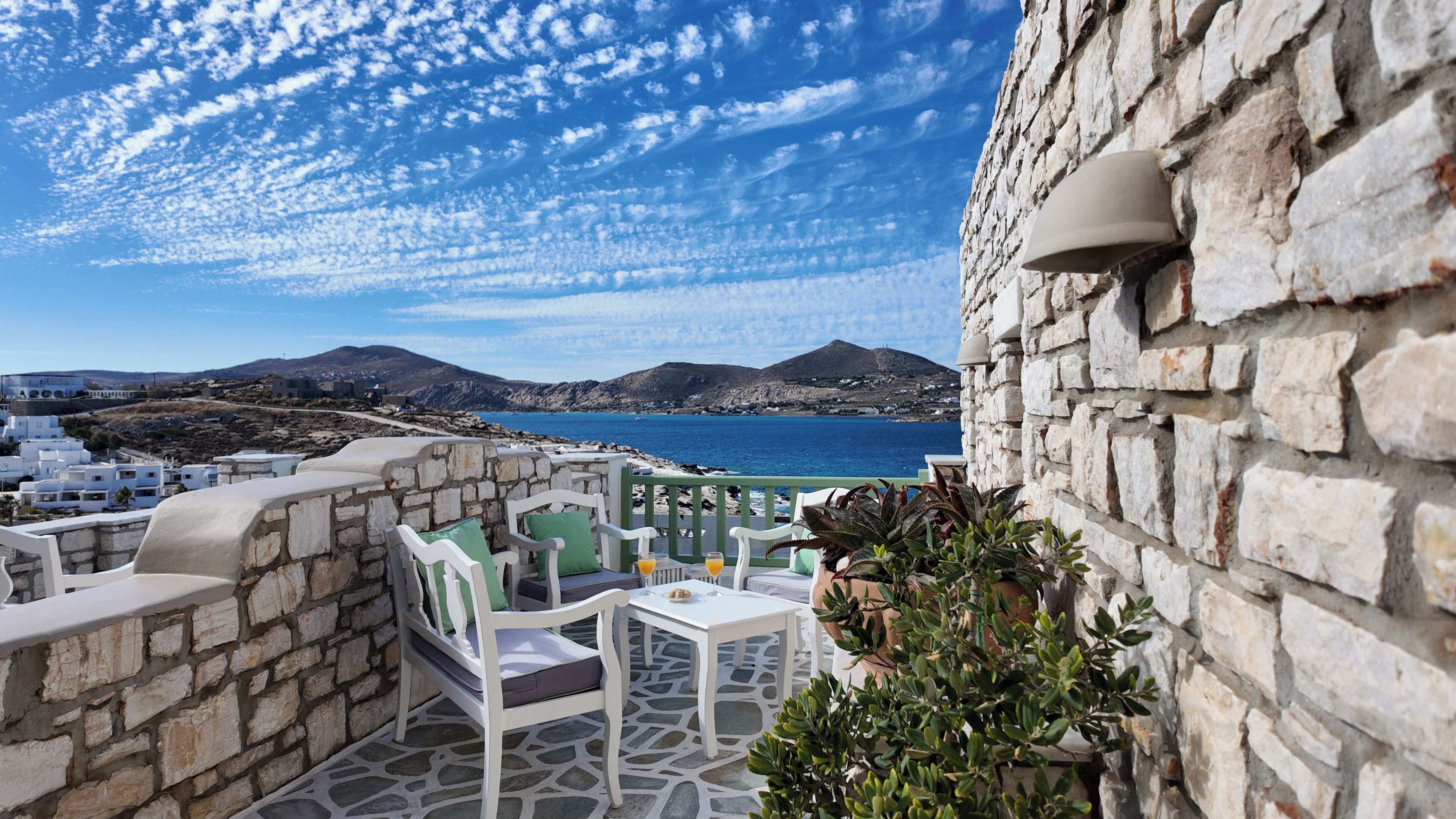 A stone terrace with white furniture, a view of the sea, and distant mountains.