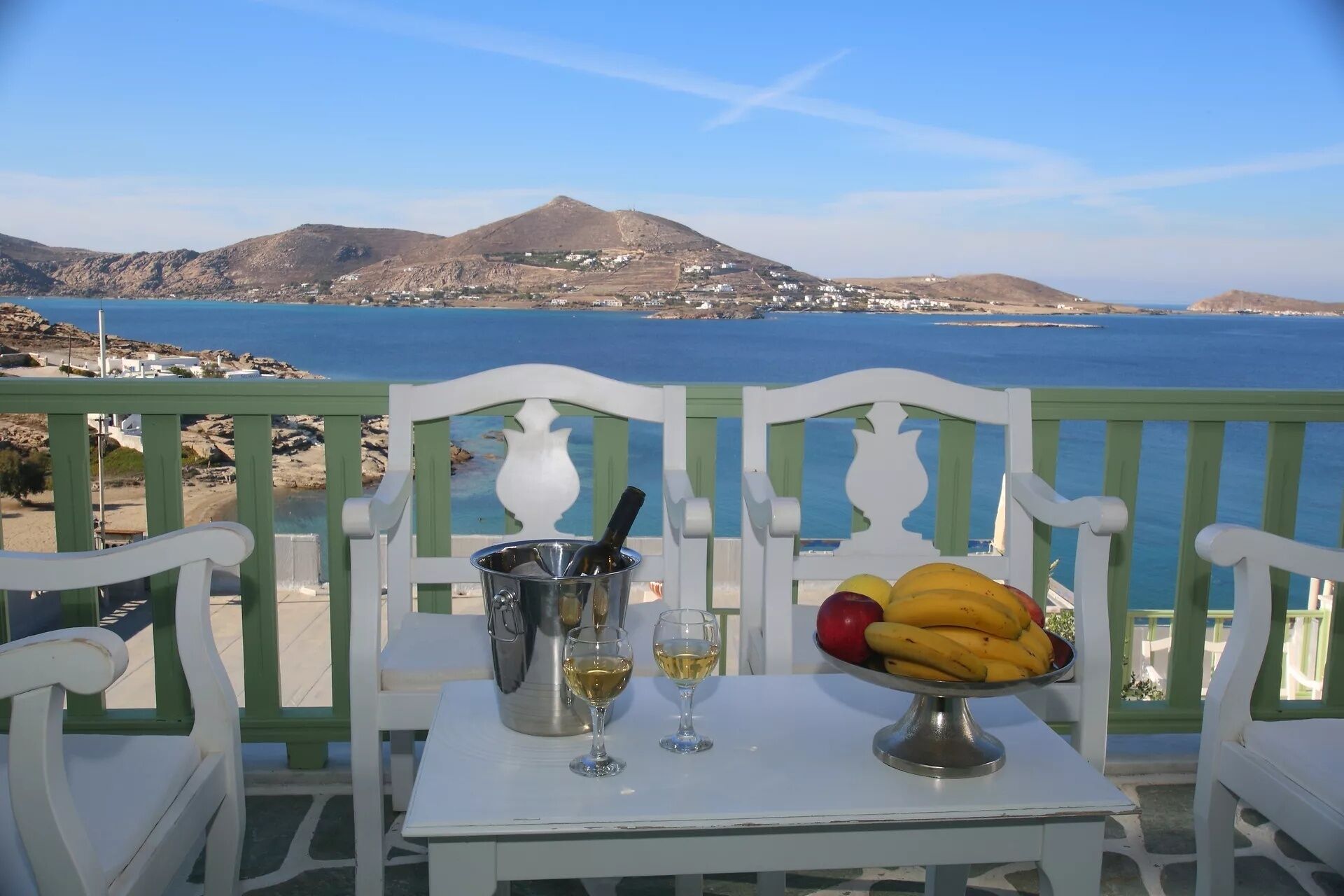 A balcony with a table set for two, featuring a bottle of wine, two glasses, and a bowl of fruit. The view includes a coastal landscape with mountains and a body of water.