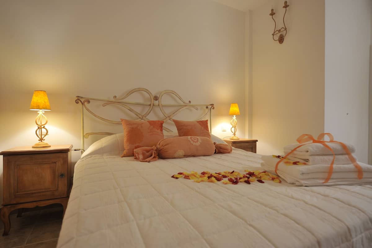 A neatly made bed with a white comforter and peach pillows, two bedside lamps, and a stack of towels with a ribbon.
