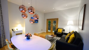 Una sala de estar moderna con una mesa redonda de comedor, sillas amarillas, un sofá negro con cojines amarillos y un televisor montado en la pared.