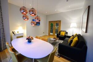 A modern living room with a round dining table, yellow chairs, a black sofa with yellow cushions, and a television mounted on the wall.