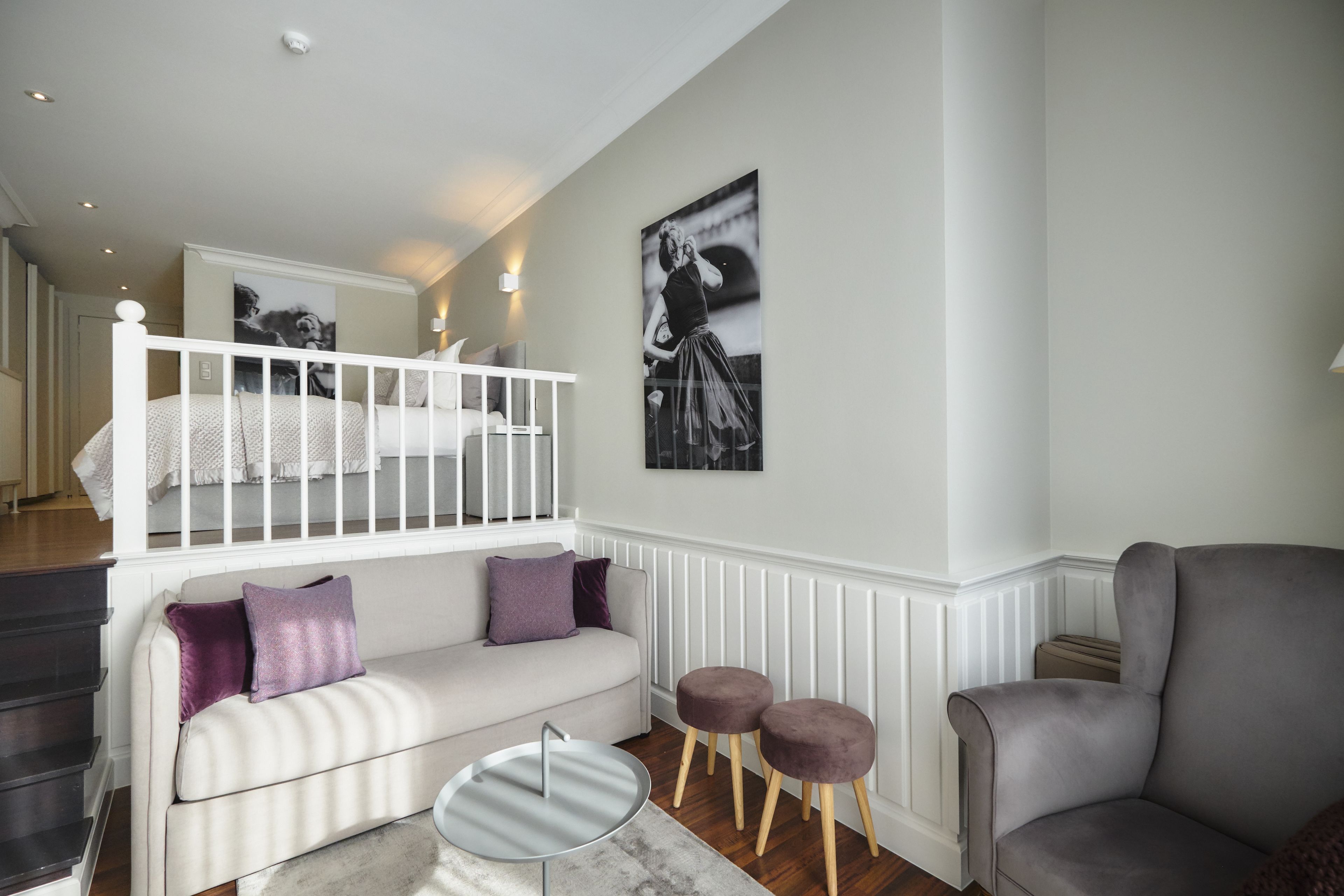 A modern living room with a white sofa, grey armchair, and a small round table. A staircase leads to an upper level.