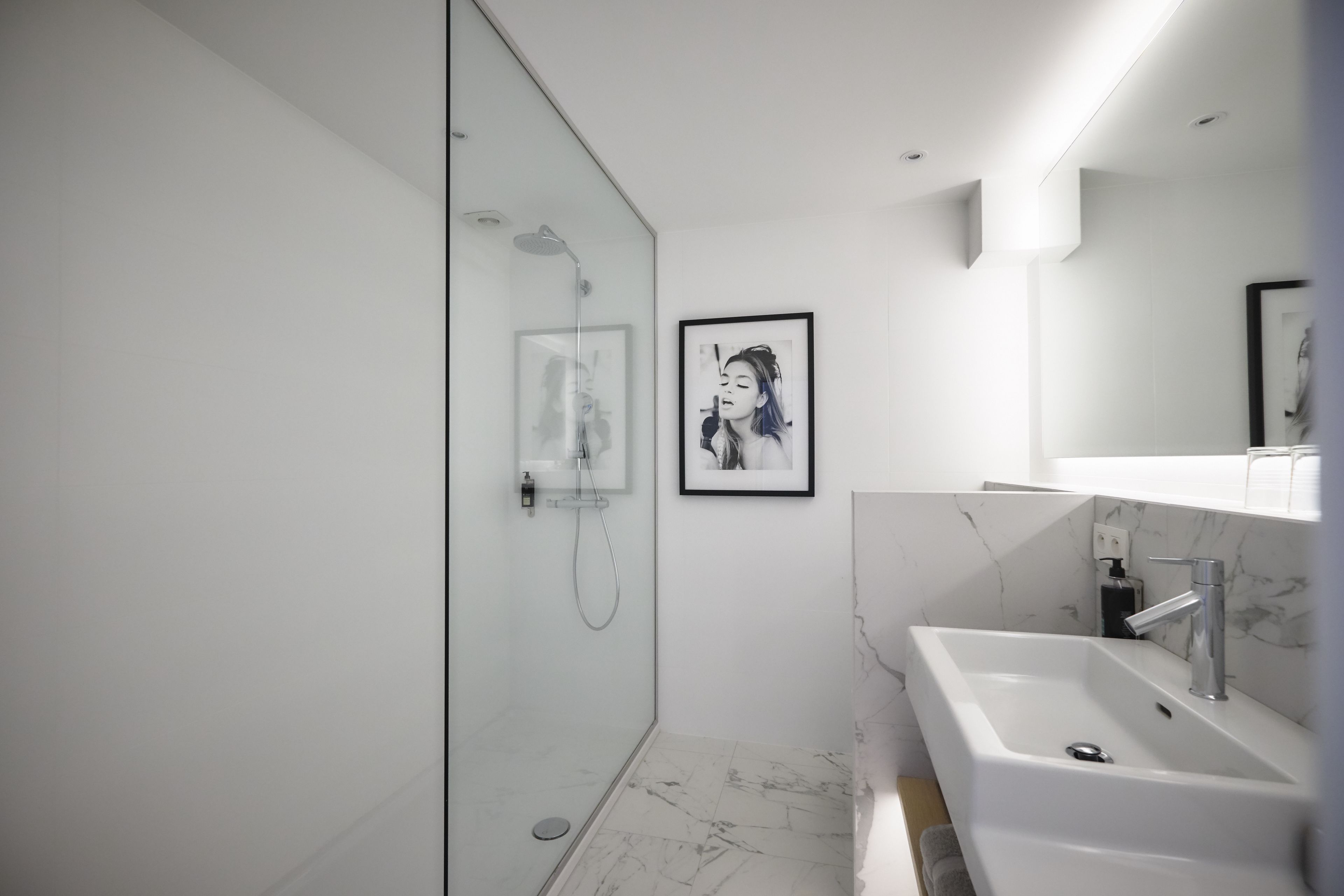A modern bathroom with a glass shower enclosure, a marble-tiled floor, a white sink with a marble countertop, and a framed picture on the wall.