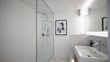 A modern bathroom with a glass shower enclosure, a marble-tiled floor, a white sink with a marble countertop, and a framed picture on the wall.
