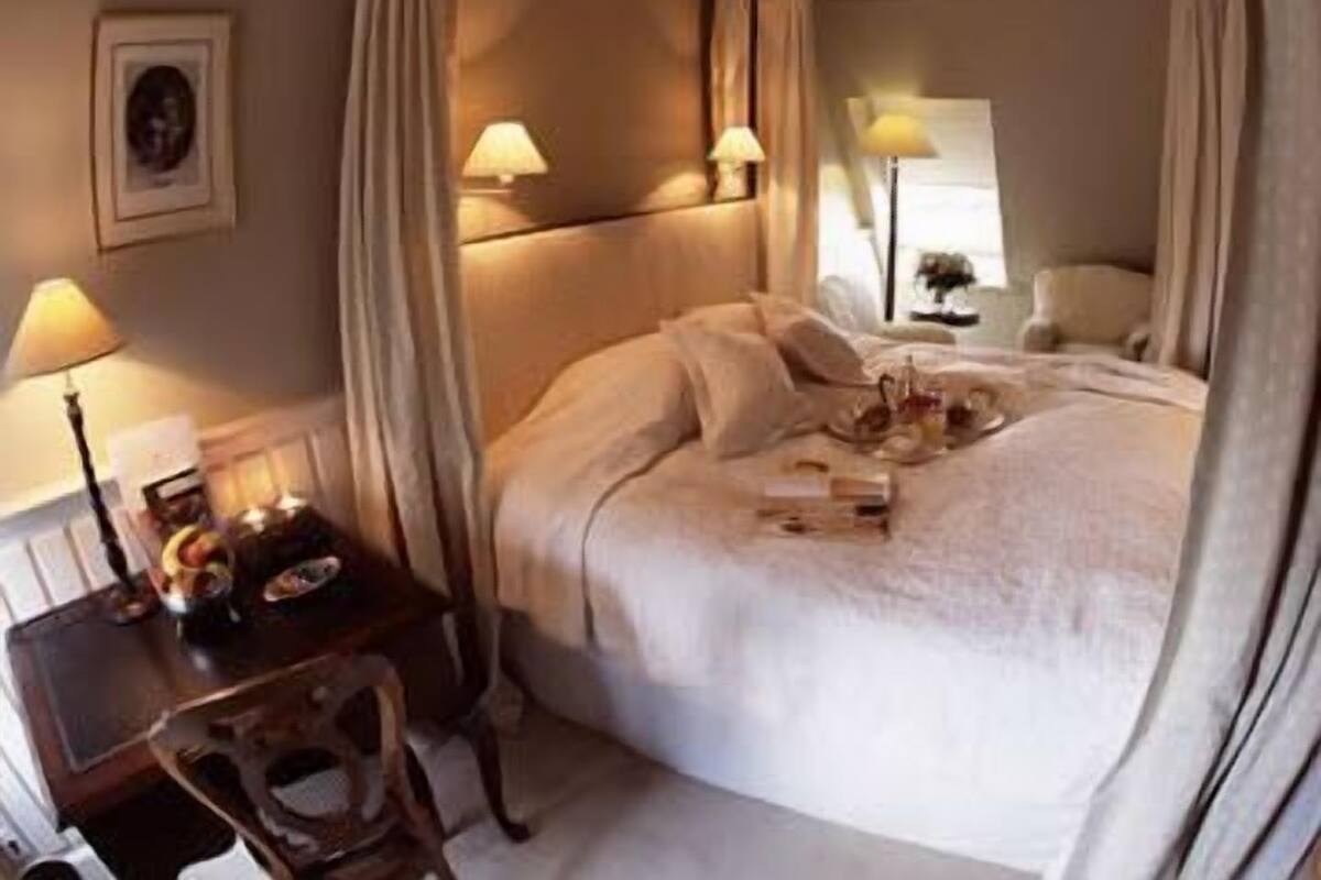 Een hemelbed met een baldakijn, bedlampen, een klein tafeltje met een dienblad en een ingelijste foto aan de muur.