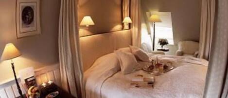 A four-poster bed with a canopy, bedside lamps, a small table with a tray of items, and a framed picture on the wall.