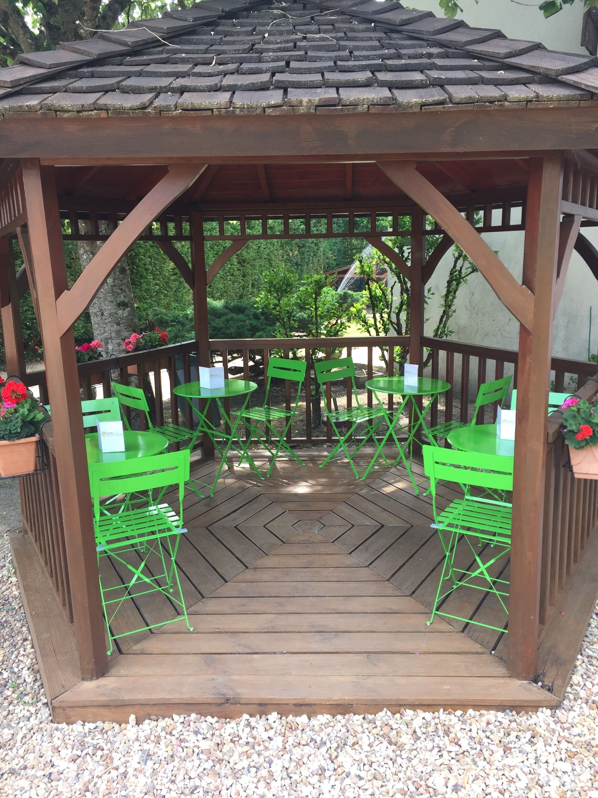 A wooden gazebo with a tiled roof, green metal chairs, and tables, surrounded by gravel and potted plants.