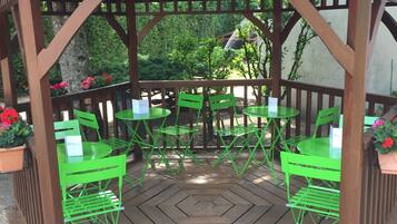 A wooden gazebo with a tiled roof, green metal chairs, and tables, surrounded by gravel and potted plants.