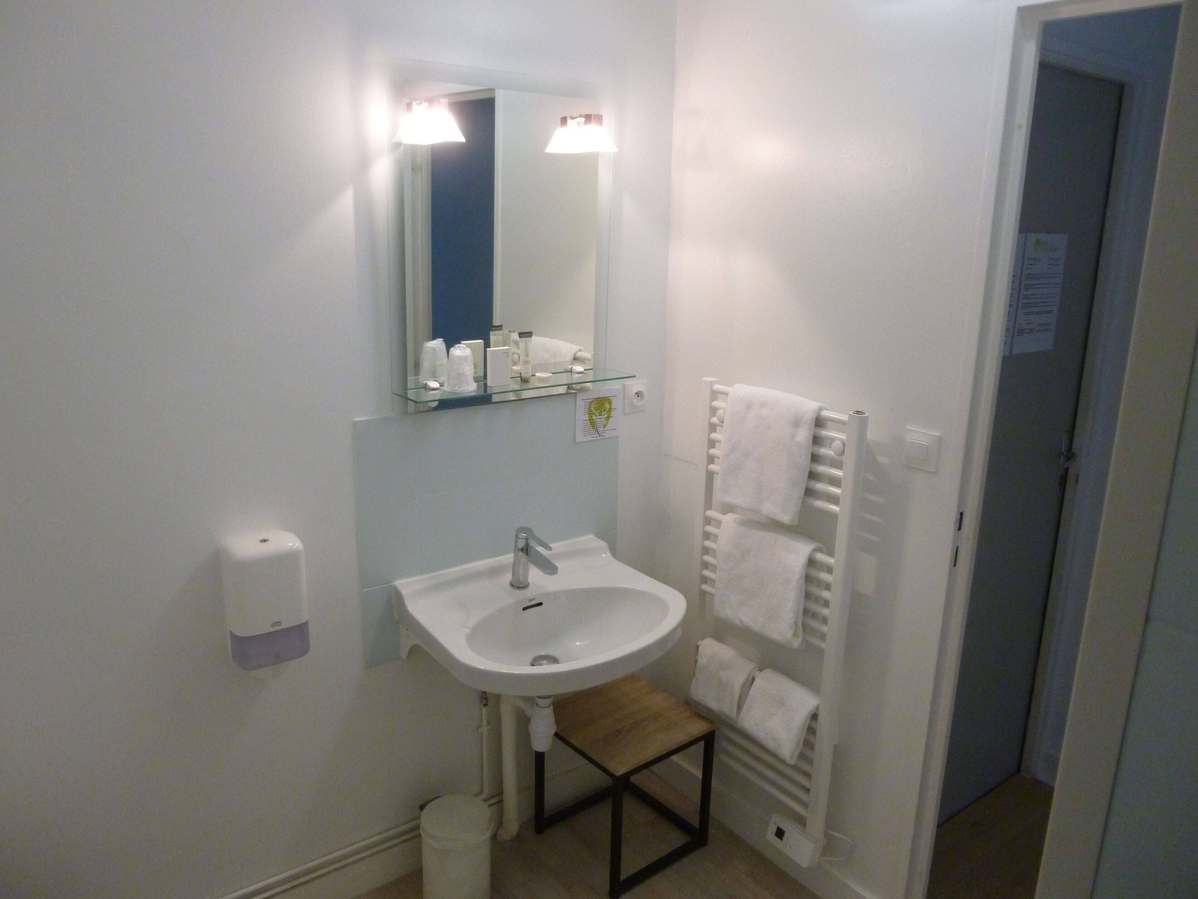 A small bathroom with a white sink, a mirror, a towel rack, and a small wooden stool.