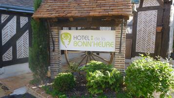 A sign for Hotel de la Bonn'Heure with a rustic design, a wooden wheel, and a garden area with plants.