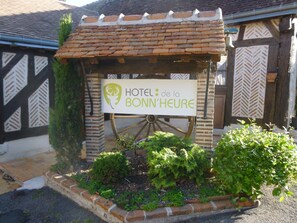 Letrero del Hotel de la Bonn'Heure con diseño rústico, una rueda de madera y un área de jardín con plantas.