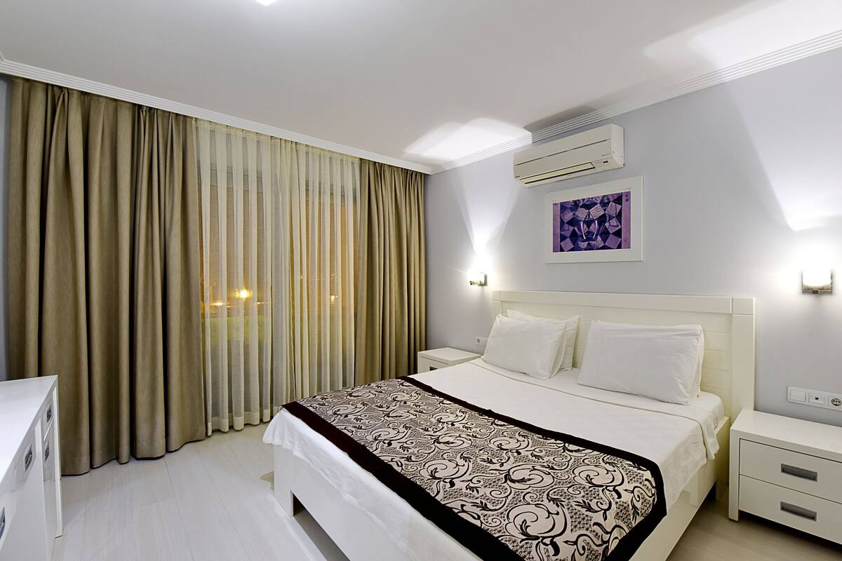 Habitación de hotel con cama, cortinas, equipo de aire acondicionado, mesita de noche y lámpara de pared.