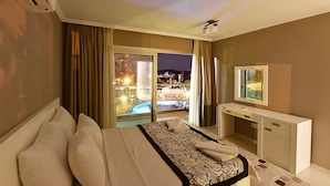 A hotel room with a bed, a desk, a TV, and a view of a pool at night. - Royal Palm Residence (Bodrum)