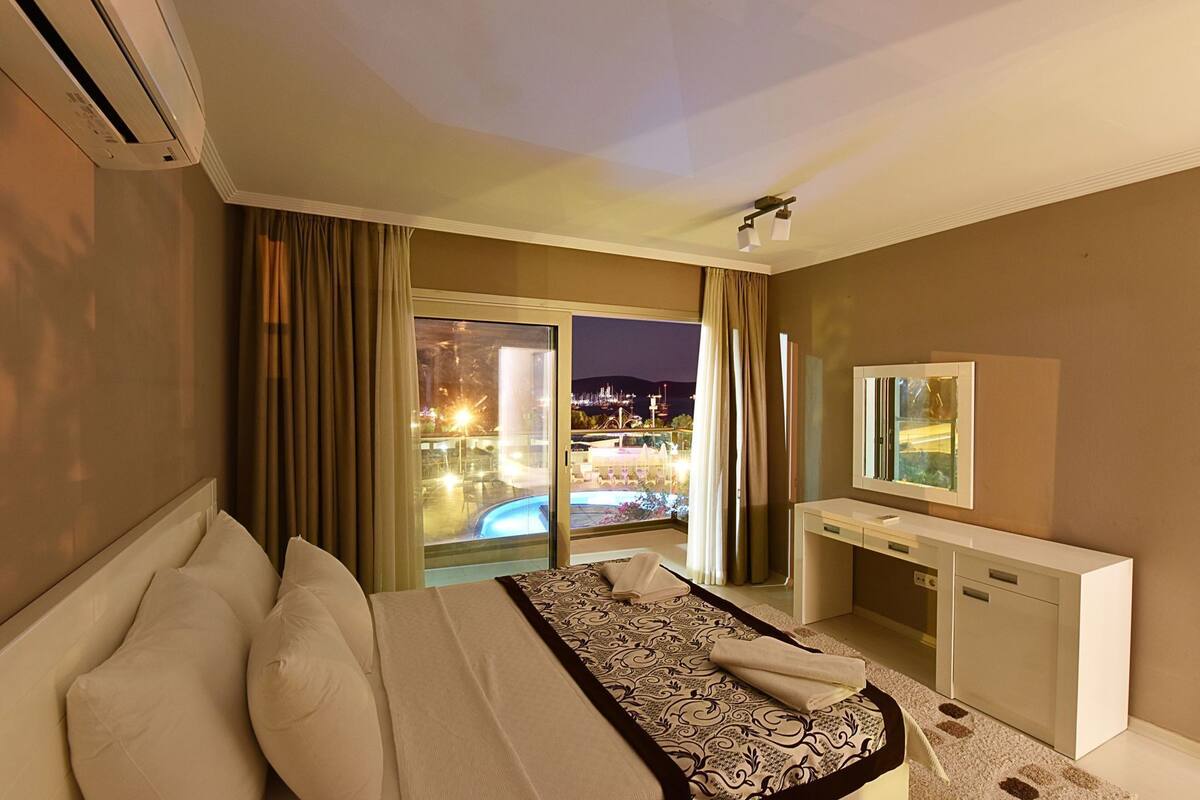 Habitación de hotel con cama, escritorio, espejo y vistas a una piscina y luces de la ciudad.