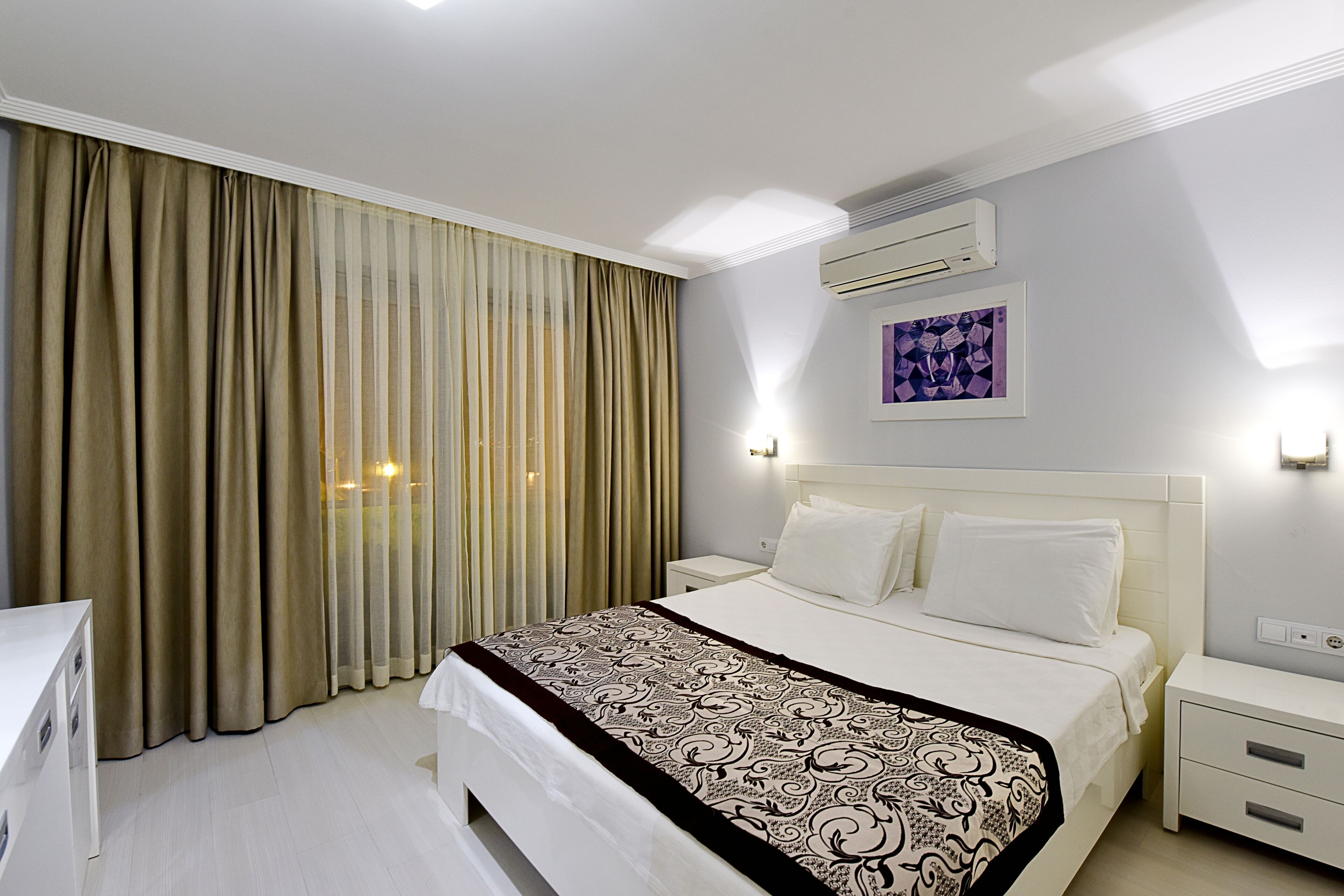 Une chambre d’hôtel avec un grand lit, une table de chevet, une commode, un bureau et une fenêtre avec des rideaux.