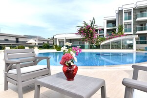 Un área junto a la piscina con un florero con forma de corazón, sillones de descanso grises y un edificio moderno al fondo.