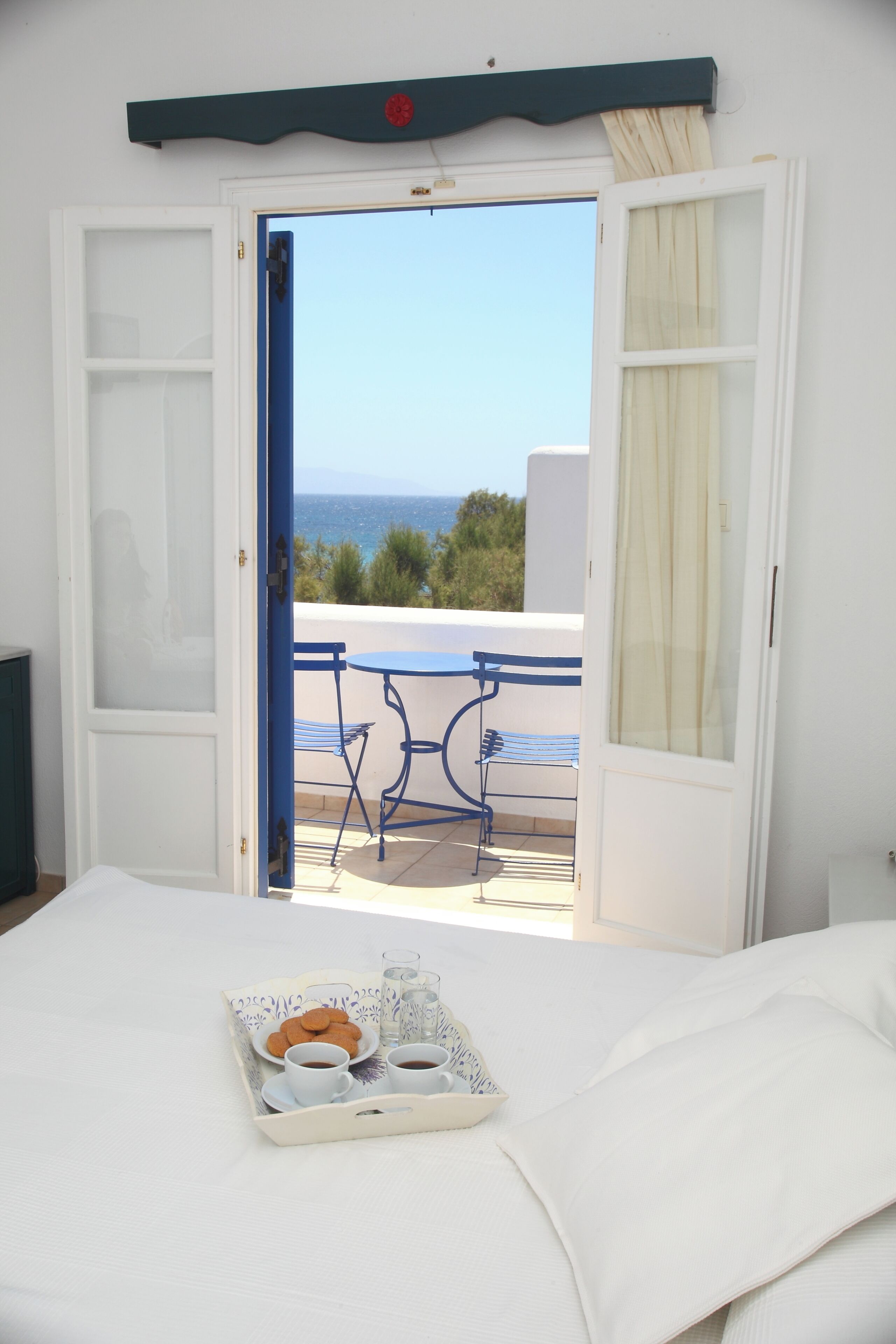 Une porte de chambre ouverte donnant sur un balcon équipé d’une table et de chaises, un plateau avec des biscuits et des tasses à café, et une vue sur la mer.